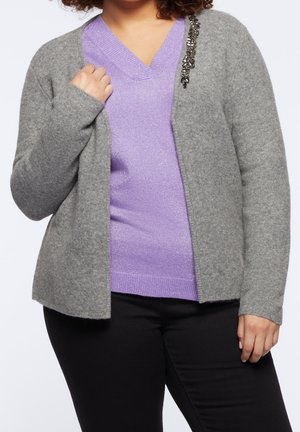 Personne portant un pull à col en V lavande sous un cardigan gris ouvert avec une broche décorative sur l'épaule gauche et un pantalon noir.