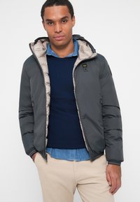Giacca a vento grigia con interno beige e cappuccio. Presenta chiusura con zip, tasche laterali e un logo sul petto. Indossata sopra un maglione blu navy e una camicia.