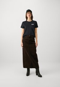 Juodas marškinėliai su baltu logo, derinami su rudos džinsinės maksi sijonu ir juodais aukštakulniais botais. Aprangą papildo juodas kepurė.