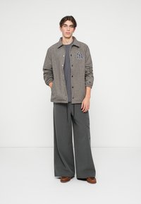 Tânăr stând cu mâinile în buzunare, purtând o jachetă cu carouri și cu logo-ul NY, un tricou gri, pantaloni largi cu dungi și pantofi maro.