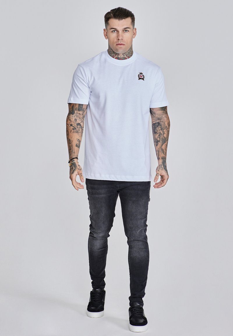 Weißes Baumwoll-T-Shirt mit rundem Ausschnitt und kurzen Ärmeln, das ein kleines gesticktes Emblem auf der linken Brust zeigt. Kombiniert mit dunklen Slim-Fit-Jeans.