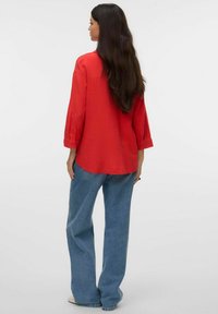 Chemise rouge à manches longues, boutonnée, avec une coupe décontractée, associée à un jean bleu ample. Texture lisse et design simple avec des manches retroussées.