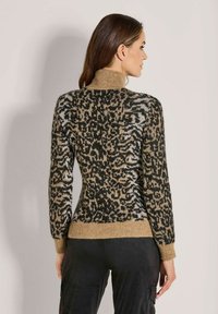 Frau mit braunem Haar, die einen hochgeschlossenen, leopardmusternden Pullover und schwarze Hosen trägt, von hinten vor einem schlichten Hintergrund fotografiert.