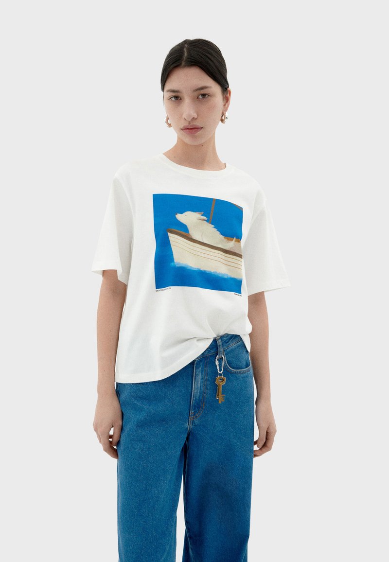 Witte katoenen T-shirt met een blauwe afbeelding van een hond op een boot. Korte mouwen, ontspannen pasvorm, gecombineerd met hoge blauwe jeans.