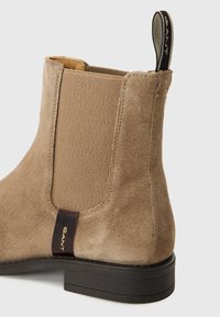 Bottine en suede beige avec des panneaux latéraux élastiques et une semelle texturée noire. Présente une étiquette de logo GANT à l'arrière pour le marquage.