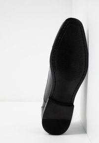 Chaussure en cuir noir avec un dessus lisse et une semelle en caoutchouc texturée. Présente un orteil arrondi et des coutures visibles le long des bords.