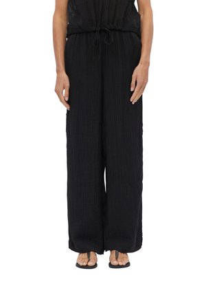 Personne portant un pantalon noir ample avec une taille à cordon et des sandales noires, debout sur un fond blanc.