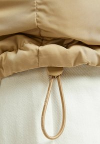 Beige jacka med huva som har en slät, glänsande yta; har en snörning för stängning och en vit, texturerad tyg under.