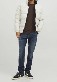 Giacca imbottita bianca con cerniera nera, maglione marrone, jeans di denim blu e sneakers nere con strisce bianche. Outfit casual con texture stratificate.