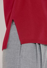 Roter Strickpullover mit seitlichem Schlitz, getragen über hellgrauen strukturierten Hosen mit sichtbarem Naht- und Taschendetail.