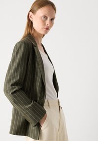 Tume roheline triibuline bleiser, millel on struktureeritud õlad, kolm nööpaugulist nööpi mansettidel ja klappkaelus, kantud valge topi ja beežide pükstega.