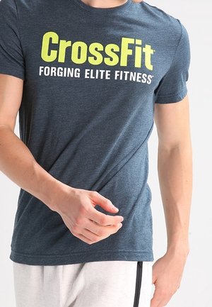 Osoba nosząca ciemnoszarą koszulkę CrossFit z napisem „FORGING ELITE FITNESS”, trzymająca koszulkę przy talii, widoczne białe spodnie.