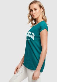 Teal bomulds t-shirt med rund halsudskæring og oprullede korte ærmer, med "BERLIN" i fede hvide bogstaver på forsiden.