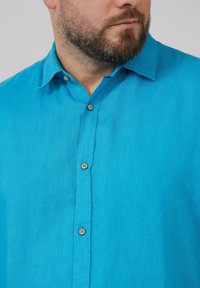 Turquoise linnen shirt met een kraag en korte mouwen. Bevat houten knopen en een geweven stof. Close-up van het voorste gedeelte.