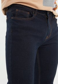 Vaqueros de un azul oscuro con corte ajustado, fabricados en denim. Presentan costuras naranjas, bolsillos frontales estándar y un diseño clásico de cinturilla.