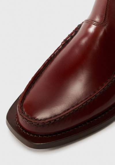 Chaussure en cuir bordeaux avec une finition lisse, accentuée par des coutures visibles le long de la tige et un bout arrondi, légèrement carré.