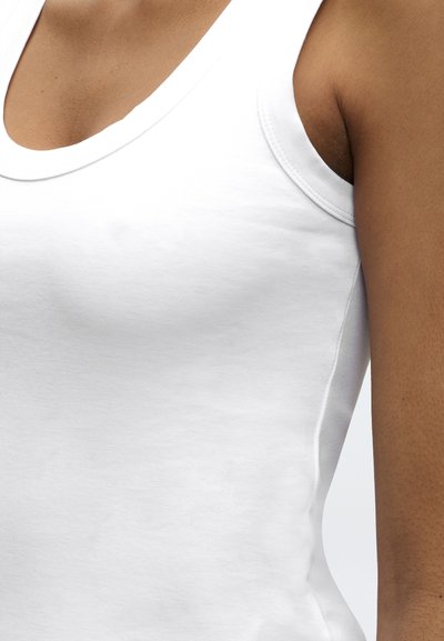 Κοντινό πλάνο ενός ατόμου που φοράει εφαρμοστή λευκή αμάνικη φανέλα, εμφανίζοντας μέρος του στήθους, του ώμου και του άνω βραχίονα.