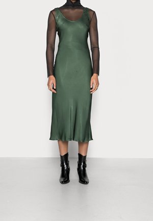 Kvinde iført en ærmeløs grøn satin midi-kjole over en gennemsigtig sort langærmet top, med sorte ankelstøvler og ringe på fingrene.