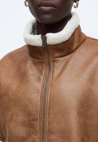 Gros plan d'une personne portant une veste en cuir marron avec un col en peau lainée blanche et des boucles d'oreilles créoles torsadées dorées.