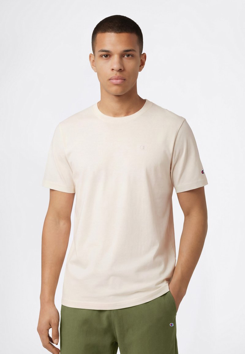 Camiseta de algodón beige claro con cuello redondo y mangas cortas, hecha de un material suave. Presenta un pequeño logo bordado en el pecho y la manga.