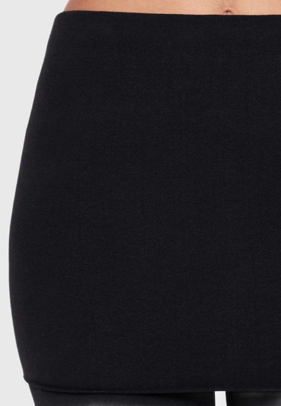 Gros plan sur une personne portant une jupe noire unie et ajustée, montrant la taille, les hanches et le haut des jambes sur un fond clair.