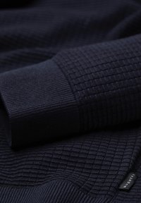 Dunkelblauer Pullover aus strukturiertem Strickstoff, mit einem karierten Muster und gerippten Bündchen. Etikett mit Markenname an der Seite sichtbar.