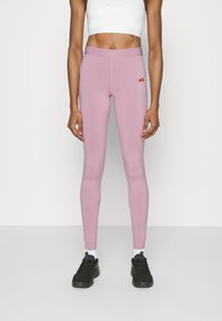 Kvinna i rosa högmidjade leggings med en liten logotyp, vit kortärmad sporttröja, vita strumpor och svarta sportskor stående mot en vit bakgrund.