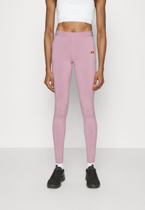 Kvinde iført pink højtaljede leggings med et lille logo, hvid cropped sports top, hvide sokker og sorte sportssko, stående mod en hvid baggrund.