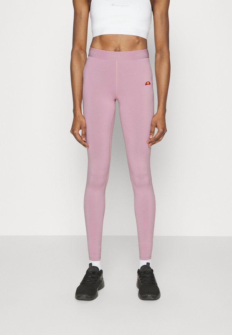 Kvinna i rosa högmidjade leggings med en liten logotyp, vit kortärmad sporttröja, vita strumpor och svarta sportskor stående mot en vit bakgrund.