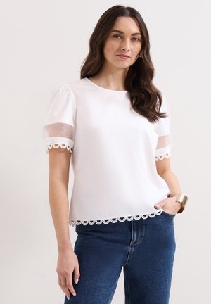 Vrouw met lang bruin haar, een witte korte mouwen blouse met kanten afwerking en blauwe spijkerbroek, staand met één hand in de zak.