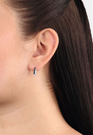 Primo piano dell'orecchio di una donna che indossa un piccolo orecchino a cerchio con gemme nere e bianche alternate.