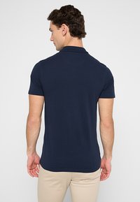 Modra polo majica iz smooth, teksturiranega blaga. Ima klasičen ovratnik in kratke rokave, s ravnim robom v pasu.