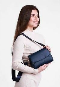 Náměsíčná modrá kožená crossbody taška s elegantním designem, magnetickým uzávěrem a nastavitelným popruhem, držená stojící osobou v neutrálním prostředí.