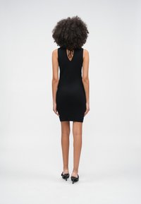Kvinna med lockigt hår som bär en ärmlös svart klänning med knytning vid övre ryggen och svarta klackskor, stående mot en vit bakgrund.