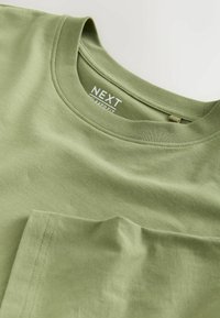 Vaaleanvihreä puuvillainen t-paita, jossa on pyöreä pääntie, rento istuvuus ja pieni taskudetaili vasemmassa rinnassa. Brändin etiketti näkyvissä.