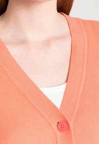 Korálový kardigan s výstřihem do V, texturovaná pletenina a jeden červený knoflík s logem na hrudi. Nositelný přes bílý top.