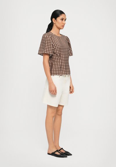 Top de cuadros marrón y blanco de manga corta, pantalones cortos blancos, zapatos negros. El top tiene un corte holgado y patrones en forma de diamante.