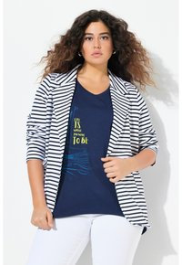 Randig kavaj i svart och vitt över en marinblå t-shirt med färgglad text. Modellen bär vita byxor, vilket visar en avslappnad, lager-på-lager-look.