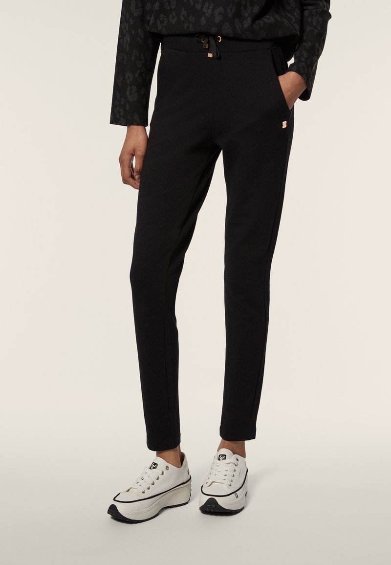 Pantaloni jogger neri realizzati in tessuto morbido, con vita regolabile con cordoncino, tasche laterali e cuciture a contrasto. Abbinati a scarpe da ginnastica bianche.