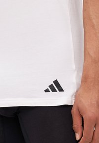Balta medvilninė marškinėlių su trumpomis rankovėmis, su juodu Adidas logotipu apatiniame kairiajame krašte. Sklandus paviršius ir reguliaraus kirpimo dizainas.