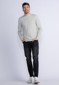 Jasnoszary sweter z fakturą i delikatnymi paskami, w połączeniu z ciemnymi dżinsami i białymi sneakerami. Model stoi z rękami w kieszeniach na jednolitym tle.