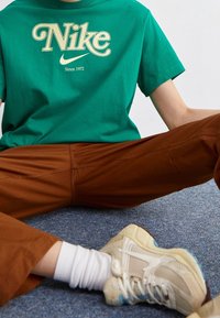 Zelené bavlněné tričko s krémovým logem Nike, hnědé kalhoty a béžové tenisky se světle modrými akcenty, sedící na šedém koberci.