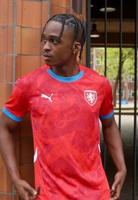 Rød Puma fodboldtrøje med et subtilt tekstureret mønster, korte ærmer og en lyseblå rund hals. Hvide logoer og våbenskjold fremhæver designet.