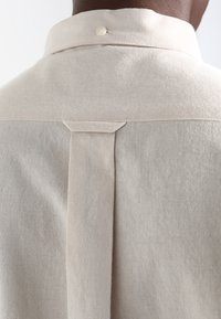 Beiges Hemd mit Kragen, das ein sauberes Rückendesign mit einer kleinen vertikalen Falte und einem einzelnen Perlenknopf-Akzent zeigt. Weicher, strukturierter Stoff.