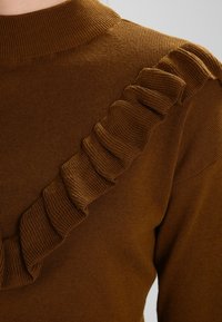Brauner Strickpullover mit einem Rüschendetail an einer Schulter, ribbeliger Textur und hohem Kragen.