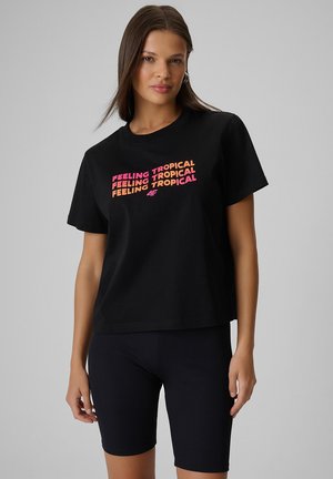 Kobieta ubrana w czarny t-shirt z napisem "FEELING TROPICAL" różowym i pomarańczowym kolorem oraz czarne prążkowane szorty rowerowe, stojąca na szarym tle.