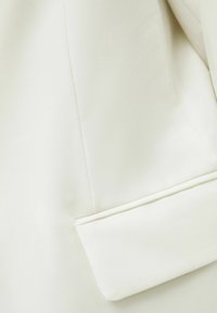 Bílá, texturovaná látka saka s klopou a precizním prošíváním podél okrajů a čistým, minimalistickým designem.