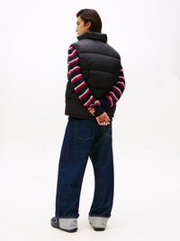 Svart pufferväst över en röd och vit randig tröja, tillsammans med lösa blå jeans och svarta skor med grå accenter.