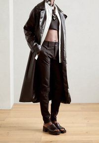 Černý kožený trenčkot přes černý crop top, vysoké černé kalhoty a robustní černé sandály na klínku, umístěné na neutrálním pozadí.