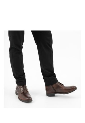 Braune Leder-Schnürstiefel, kombiniert mit schwarzen Hosen vor weißem Hintergrund.